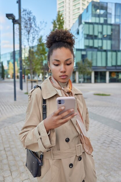 une femme vêtue d'un manteau porte un sac tient un téléphone portable fait défiler les pages Web lit les nouvelles en ligne regarde attentivement l'écran du téléphone portable aime la communication en ligne fait du shopping