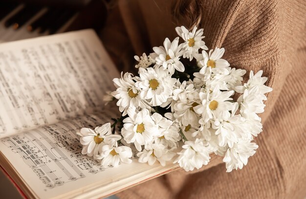 Une femme vêtue d'un pull douillet tient une collection de notes et un bouquet de fleurs, assise près du piano.