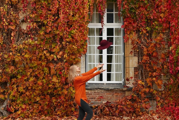 Une femme vêtue d'un pull orange jette son chapeau près d'un mur avec des raisins sauvages en automne