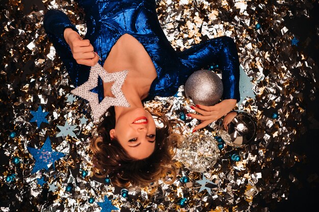 Une femme vêtue d'une robe bleue à paillettes sourit et s'allonge sur le sol sous une chute de confettis multicolores.