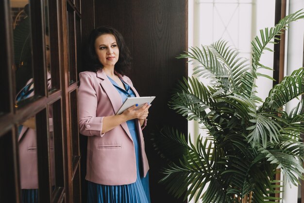 Une femme vêtue d'une robe bleue et d'une veste rose se tient près de la porte du bureau en souriant tenant une tablette