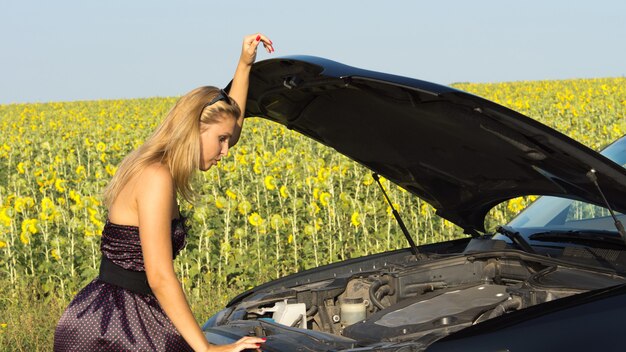 Femme vêtue d'une robe élégante vérifiant sous le capot surélevé de son véhicule alors qu'elle tente de déterminer la raison de la panne