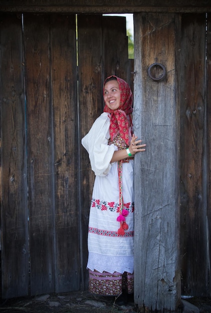 Une femme vêtue d'une robe folklorique russe et d'une écharpe debout à la porte en bois et souriante