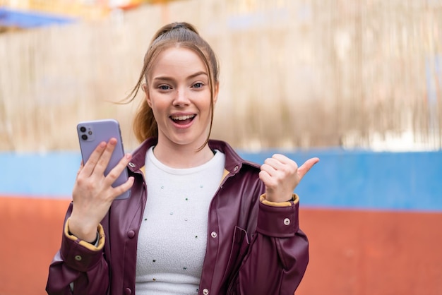 Une femme vêtue d'une veste en cuir violet tient un téléphone et montre un pouce levé.