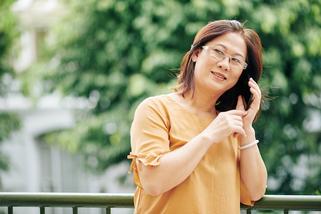 Femme vietnamienne mature dans des verres debout à l'extérieur et parlant au téléphone avec un parent ou un ami