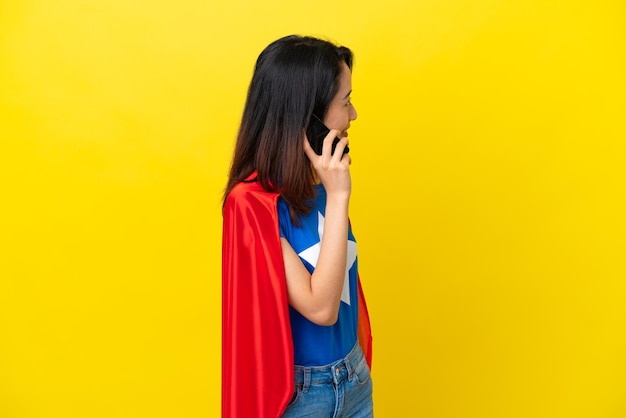 Femme vietnamienne de super héros isolée sur fond jaune gardant une conversation avec le téléphone portable avec quelqu'un