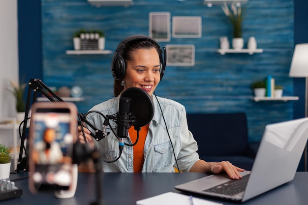 Femme vlogger avec un casque regardant un ordinateur portable et enregistrant une vidéo. Influenceur diffusant un podcast en ligne sur smartphone. Créateur de contenu en home studio utilisant un équipement de diffusion audio.