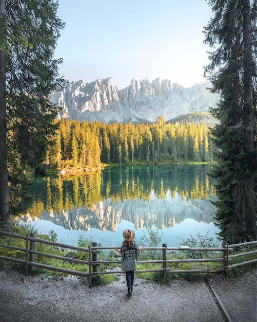 Une femme voagere dans un chapeau observe un lac avec des arbres et des montagnes se reflétant dans le lac Si Carezza