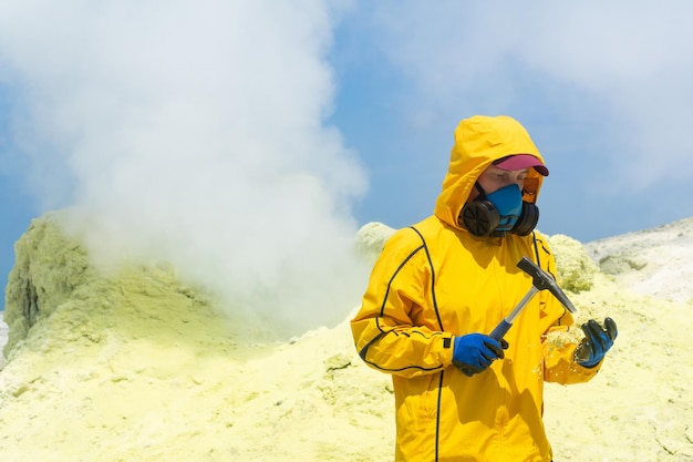 Femme volcanologue sur le fond d'une fumerolle fumante examine un échantillon d'un minerai de soufre avec un marteau géologique