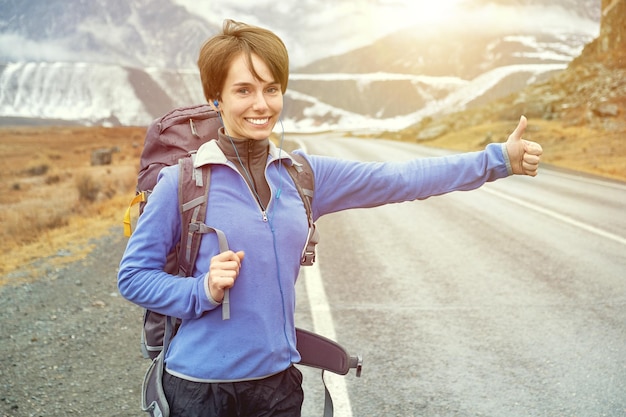 Femme de voyage faisant de l'auto-stop Belle jeune femme auto-stoppeuse par la route pendant un voyage de vacances dans les montagnes au coucher du soleil