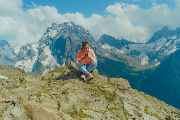 Femme voyageant à l'aide d'un smartphone dans les montagnes Femme au corps entier assise sur un rocher et naviguant sur un smartphone contre un ciel nuageux par une journée ensoleillée dans les montagnes