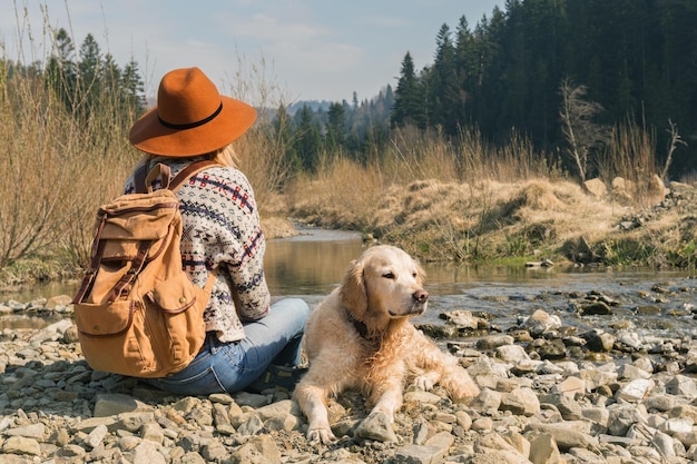 Femme voyageant et chien golden retriever dans la nature Hipster élégant en chapeau marron et pull en laine avec sac à dos en textile vintage près de la rivière des montagnes et de la forêt Concept de voyage et d'aventure