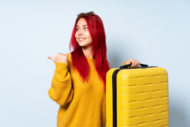 Femme voyageur tenant une valise isolée sur mur bleu pointant vers le côté pour présenter un produit