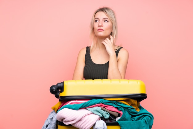 Femme voyageur avec une valise pleine de vêtements sur le mur rose isolé en pensant à une idée