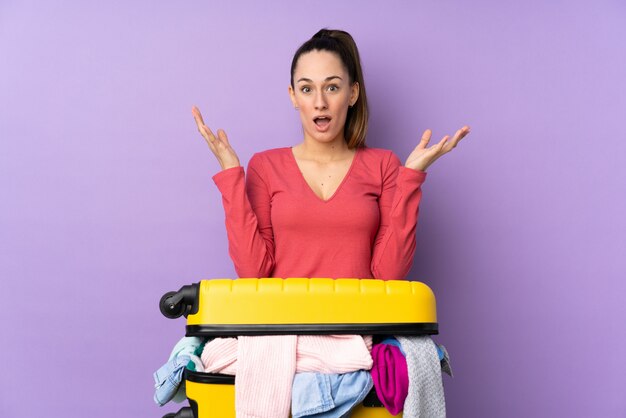 Femme voyageur avec une valise pleine de vêtements sur un mur violet isolé avec une expression faciale surprise