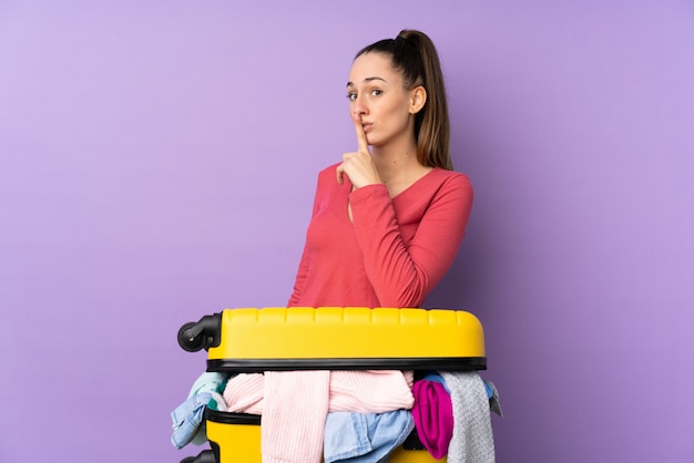 Femme voyageur avec une valise pleine de vêtements sur un mur violet isolé faisant un geste de silence