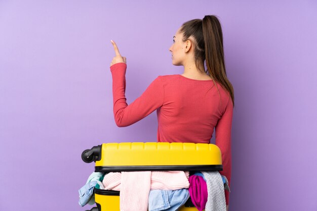 Femme voyageur avec une valise pleine de vêtements sur un mur violet isolé pointant vers l'arrière avec l'index