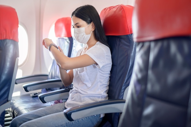 Une femme voyageuse portant un masque de protection à bord de l'avion à l'aide d'une montre intelligente, voyage sous la pandémie de Covid-19, voyages de sécurité, protocole de distance sociale, nouveau concept de voyage normal
