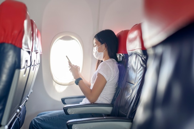 Une Femme Voyageuse Portant Un Masque De Protection à Bord De L'avion à L'aide D'un Smartphone, Voyage Sous La Pandémie Covid-19, Voyages De Sécurité, Protocole De Distance Sociale, Nouveau Concept De Voyage Normal