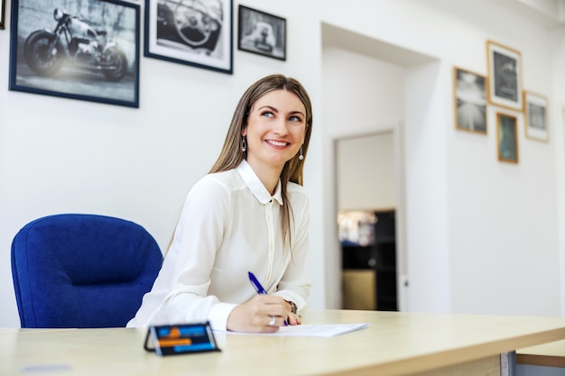 Femmes d'affaires dans le bureau d'inspection technique. Une femme adulte est assise à une table devant un mur blanc et tient un crayon à la main tout en signant des papiers. Service de voiture, signature de la documentation