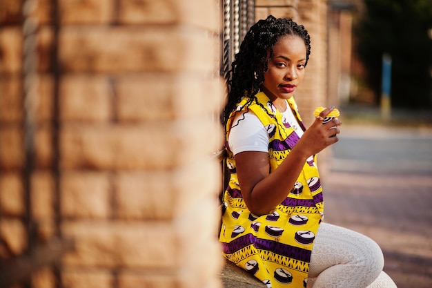 Femmes afro-américaines élégantes en veste jaune posées dans la rue avec une boisson chaude dans une tasse en papier jetable