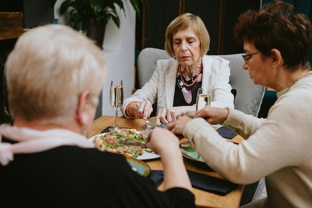 Femmes âgées blondes et brunes vêtues de noir et blanc assises à table, mangeant de la pizza avec des couteaux et des fourchettes, buvant du vin ou du champagne tout en célébrant leur réunion ou des vacances