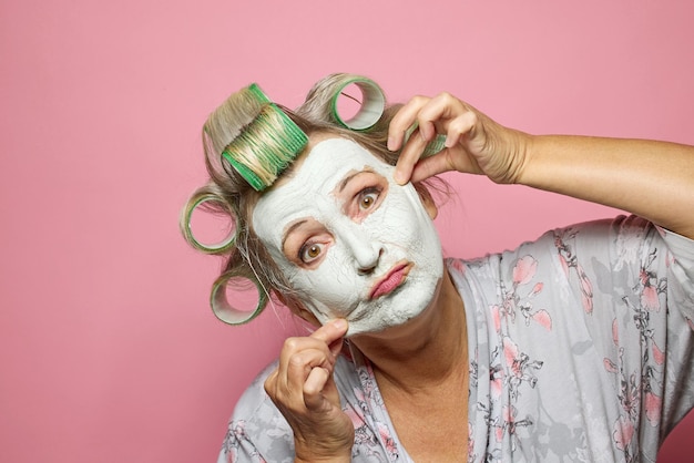 Photo des femmes âgées drôles avec un masque en argile et des rouleaux de cheveux sur un fond rose