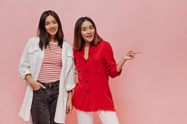 Les femmes asiatiques bronzées aux yeux bruns dans des chemises surdimensionnées sourient et regardent la caméra sur une jeune fille isolée en chemisier rouge pointe à la place du texte sur fond rose