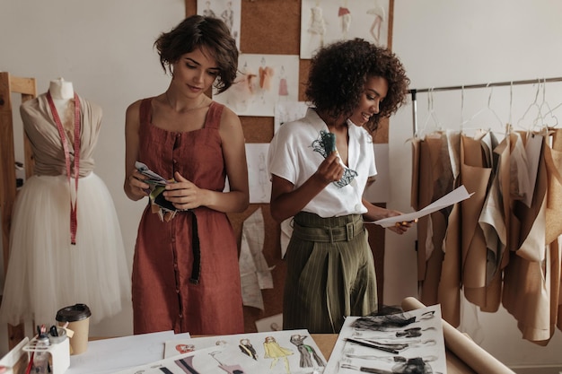 Des femmes attirantes dans des tenues à la mode tiennent des échantillons de dentelle et de textile Les créateurs de mode travaillent dans leur bureau confortable et lumineux