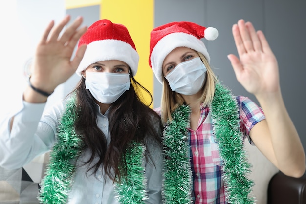 Femmes en chapeaux de Noël rouges et masques médicaux sur leurs visages en agitant bonjour