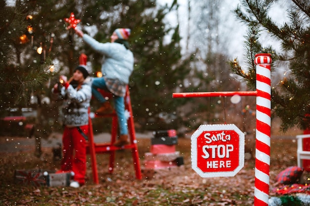 Les femmes décorent un arbre de Noël à l'extérieur, un cierge magique allume un panneau d'arrêt avec le texte Santa stop here.