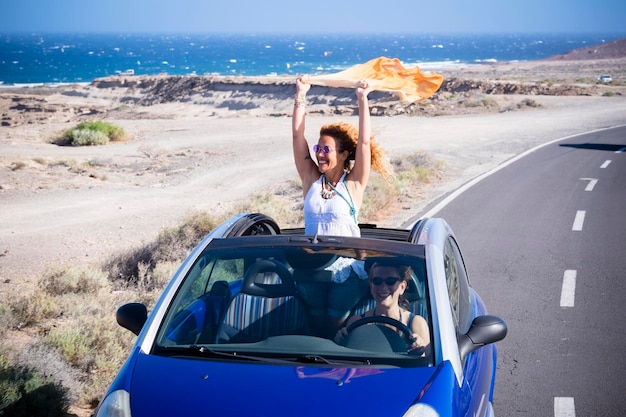 Des femmes heureuses voyagent en voiture avec une voiture décapotable Une femme à l'extérieur du toit s'amuse Destination pittoresque du désert Conduite automobile Couple d'amis excités dans un véhicule en voyage Aventure