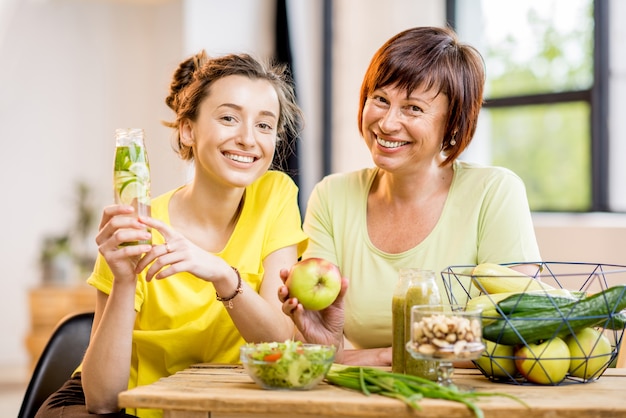 Femmes jeunes et plus âgées assises avec des aliments sains et des boissons fraîches après l'entraînement sportif à l'intérieur sur fond de fenêtre