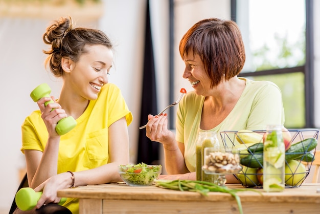 Femmes jeunes et plus âgées assises avec des aliments sains et des boissons fraîches après l'entraînement sportif à l'intérieur sur fond de fenêtre