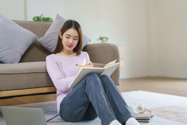 Femmes lisant des données commerciales sur un ordinateur portable et écrivant des notes tout en étant assises sur le sol dans leur style de vie à la maison