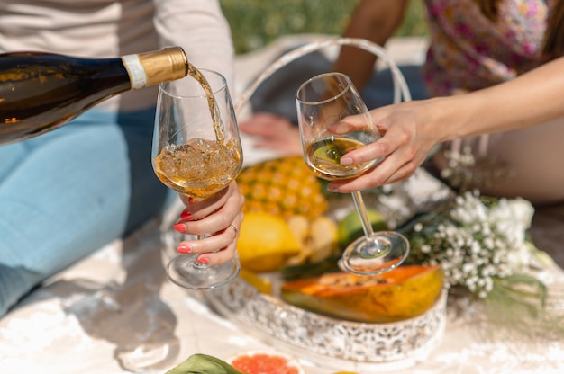 Femmes méconnaissables assises sur une couverture en train de pique-niquer. Femme versant du vin blanc dans un verre. Fruits tropicaux sur fond