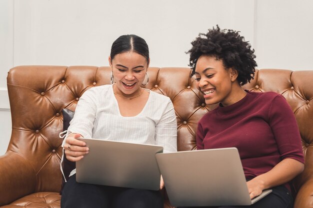 Les femmes multiculturelles à l'aide d'un ordinateur portable sur un canapé se détendre sur le salon et rire de quelque chose à l'écran