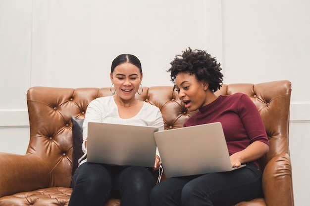 Femmes multiculturelles à l'aide d'un ordinateur portable sur un canapé se détendre sur le salon et rire de quelque chose sur l'écran