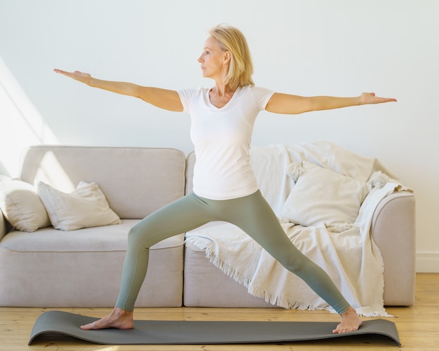 Femmes mûres à la retraite ciblées en vêtements de sport pratiquant la pose de yoga guerrier à la maison