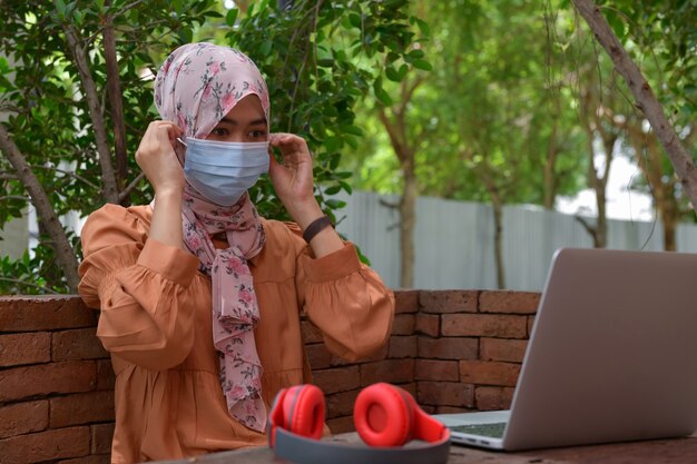Les femmes musulmanes portent des masques et utilisent des écouteurs pour écouter de la musique sur un ordinateur portable. Pour se détendre pendant la 19e épidémie de covid, le concept de garder la distance sociale