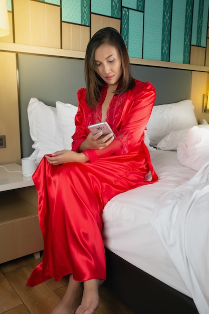 Les femmes qui travaillent ont mis une alarme pour se réveiller sur un téléphone portable dans la chambre du complexe. Fille en robes de satin rouge à l'aide d'un téléphone portable assis sur un lit blanc