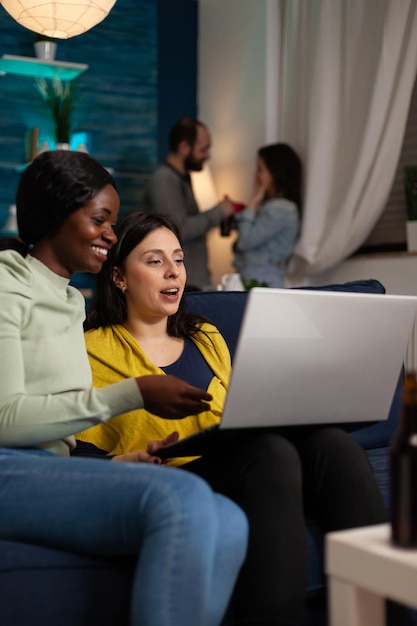 Femmes souriantes regardant une vidéo amusante sur Internet tenant un ordinateur portable assis sur un canapé et profitant de passer du temps ensemble. Groupe de personnes traînant pendant la fête du week-end à la maison. Notion d'amitié