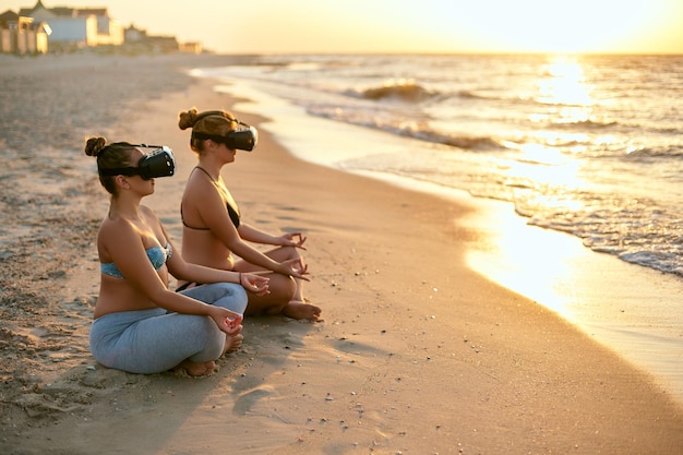 Les femmes utilisent des lunettes vr pour une immersion plus profonde deux femmes faisant de la méditation de yoga en groupe sur la plage en