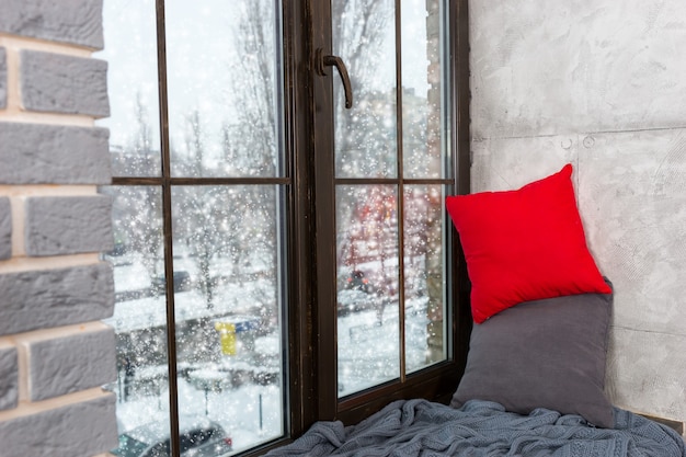 Fenêtre avec rebord de fenêtre avec oreillers et couverture dans la chambre de style loft, à l'extérieur de la fenêtre neige