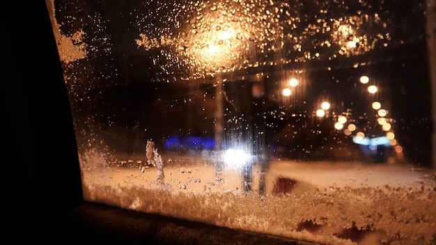 Fenêtre de voiture embué La fonte des neiges sur une fenêtre de voiture Phares de voiture bokeh et reflets de couleur vive derrière le verre enneigé