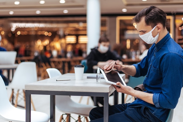 Fermer . homme dans un masque de protection essuyant l'écran d'une tablette numérique avec un antiseptique.