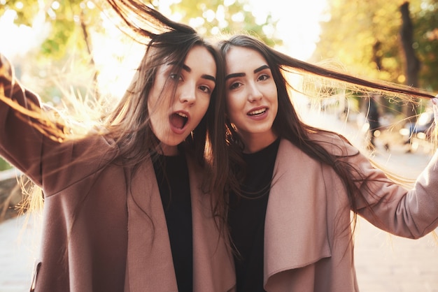 Fermer le portrait de deux jolies et gaies, jeunes filles jumelles brune en manteau décontracté s'amuser et jouer avec les cheveux en plein air dans un parc d'automne ensoleillé sur l'arrière-plan flou.