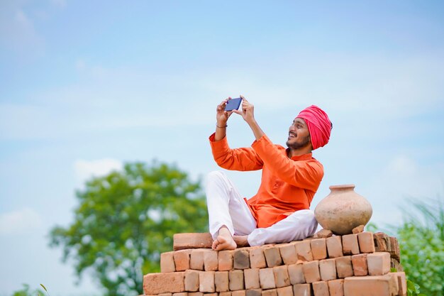 Fermier indien assis sur des briques et utilisant un smartphone sur le terrain agricole.