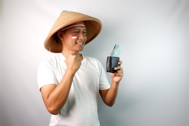 Un fermier indonésien portant un chapeau conique montre un visage excité tout en tenant son portefeuille plein d'argent