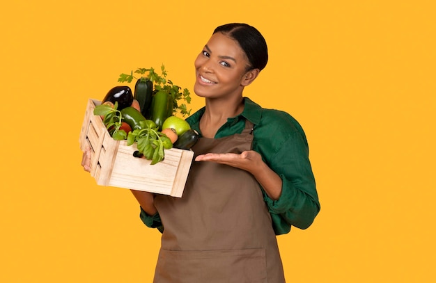 Une fermière africaine montrant des légumes sur fond jaune.